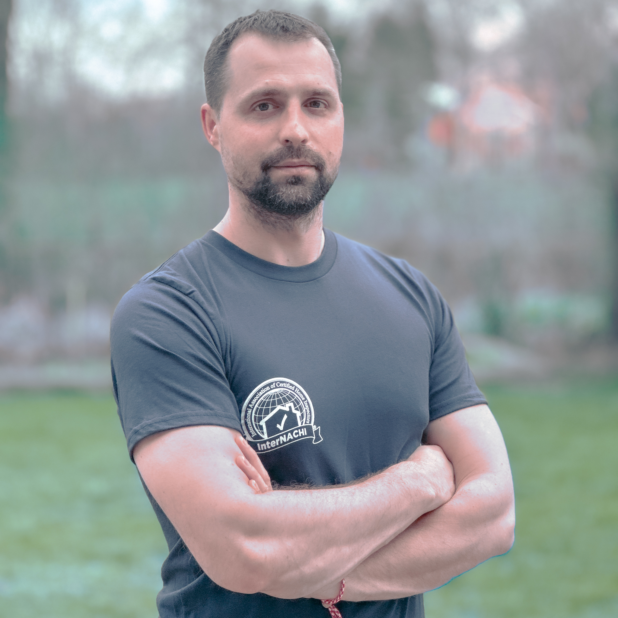 Valeri Georgiev, known as Val, stands confidently with his arms crossed. He's wearing a black T-shirt with the InterNACHI logo, symbolizing his certified expertise as a home inspector. Behind him is a soft-focus view of a residential area, reflecting his real estate industry background and property investment experience. His expression is serious and professional, indicative of his meticulous approach to home inspections and commitment to providing thorough service with the latest techniques and technology.